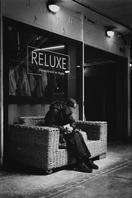A man wearing a cap and dark clothing is seated on a wicker sofa inside a shopping mall, resting and looking down at his phone or a small object in his hands. Next to him on the sofa is a black suitcase or travel bag. Behind him, there is a large storefront window with the word 'RELUXE' in bold letters and the tagline 'Uncomplicated yet elegant' visible beneath it. The store displays hanging clothing items and woven baskets inside, with the window framed in dark material. The lighting in the area is moderate, with ceiling lights illuminating the scene and casting soft shadows on the floor, which is made of polished material. The surroundings suggest a quiet moment during a home relocation or personal outing, with the seating area and shopping environment providing context for moving or packing activities related to a house or furniture transport, consistent with services offered by Man and Van Ilford.