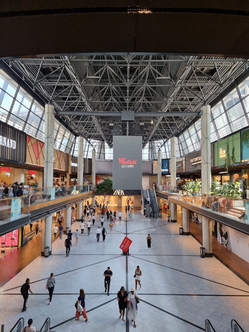 Inside the atrium of Eden Shopping Centre Ilford, the spacious area features a high glass ceiling supported by a steel framework, allowing natural light to illuminate the environment. The ground level demonstrates a large open space with numerous shoppers walking across the polished floor, some carrying shopping bags, while others are engaged in conversations. Along the upper level, a series of retail storefronts with illuminated signage, including brands such as Zara and others, line the perimeter, with glass balustrades separating the walkways from the open central area below. An escalator on the right side provides access between floors, and a prominent digital advertising screen displays the Wendy’s logo with the slogan, 'LEAD THE TRENDS.' The scene captures the bustling movement typical of a busy retail shopping environment, where furniture, boxes, and packing materials are absent, but the structure and shopping activity reflect the process of home relocation and furniture transport preparations, as coordinated by services like Man and Van Ilford.