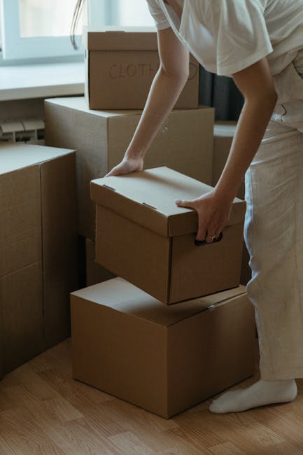 A person wearing light-colored clothing is carefully lifting a medium-sized cardboard box, marked with the word 'CLOTHES' written in red, in a room with natural light coming through a large window. Several other cardboard boxes of various sizes, some sealed with packing tape, are stacked nearby on the hardwood floor. The scene depicts an indoor home relocation process, with the individual preparing boxes for transportation as part of the packing and moving activities involved in house removals. In the background, additional boxes are visible, some on the floor and others on a piece of furniture, indicating an organized packing effort typical of professional removals services. The presence of the boxes and the action of lifting suggest an ongoing furniture transport or packing and moving operation, aligned with services offered by Man and Van Ilford for house relocations on Cranbrook Road, ILFORD.