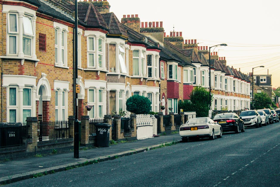 A row of terraced houses on Cranbrook Road in Ilford, featuring brick facades, bay windows with white frames, and small front gardens enclosed by low brick walls and black metal fencing. Some houses have window boxes with plants or flowers. The street is lined with parked cars along the curb, including sedans and hatchbacks in various colors. A black wheelie bin is situated on the sidewalk near the fence of one house. Street lamps and overhead electrical wires are visible, along with a clear sky indicating early evening or late afternoon lighting. The image captures the residential street environment typical of home relocation or furniture transport scenarios, where moving companies like Man and Van Ilford manage the loading process of packed boxes and furniture onto their vehicle, possibly seen beyond the frame, preparing for a house removal or move. This scene illustrates the typical street setting for house removals on Cranbrook Road in Ilford, relevant to packing, loading, and transportation logistics.