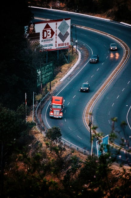 A winding dual carriageway with multiple lanes curves through a hilly area, with a red and white van parked on the roadside near a small tree and a metal guardrail. Several cars are driving along the 