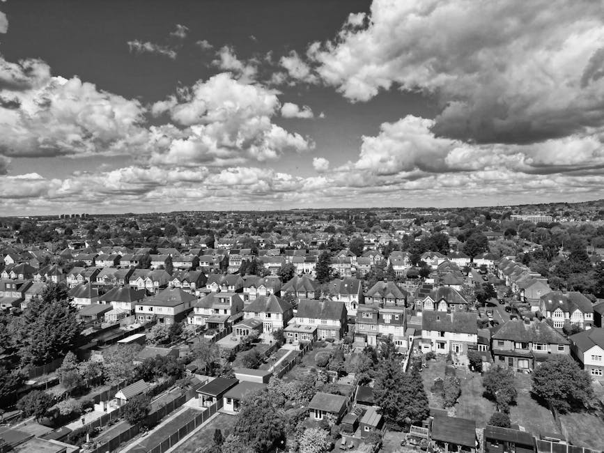 A black-and-white aerial photograph of a suburban residential area showing a dense array of houses with pitched roofs, some with small gardens and backyards separated by fences or hedges. The houses a