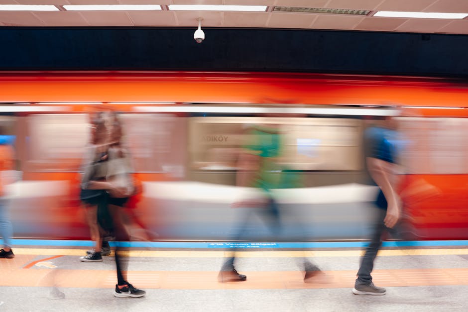 Inside Ilford station, a moving train with a bright orange exterior is captured in motion blur, with several blurred passengers visible on the platform, some standing near the edge and others walking or waiting. The platform is illuminated with bright lighting, and safety markings are visible along the edge. The image highlights the busy environment typical of a train station, which is a common location involved in house removals and transport logistics. The presence of people and the train's speed suggest the importance of efficient planning when scheduling home relocation services, including packing, loading, and transportation, as offered by Man and Van Ilford, especially when coordinating with busy transit hubs like Ilford Seven Kings station.