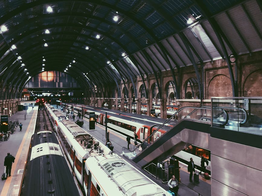 Interior view of Ilford Seven Kings station at night, showing a large arched steel roof with numerous indoor ceiling lights illuminating the platform area. The station features multiple platforms with trains parked alongside, including modern commuter trains with white and red carriages. Several passengers with luggage are walking on the platforms, some waiting near the train doors, indicating active use of the station for home relocation or furniture transport. The brick wall detailing and glass barriers at the stairs highlight the station's structural features. In the foreground, an escalator or stairway with glass panels leads to an upper level, while the platform area has digital information screens and safety signage. Bright lighting contrasts with the darker arched ceiling, emphasizing the station's architectural design suitable for efficient moving logistics, as supported by Man and Van Ilford in their removals and moving services.
