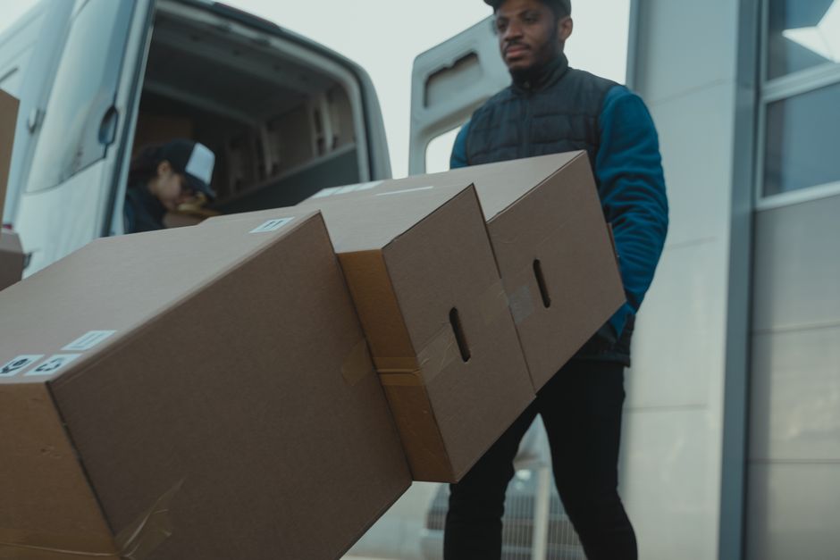 A man wearing a dark padded vest and blue long-sleeve shirt is standing outside near a white van, holding three cardboard boxes stacked together. The boxes are sealed with packing tape and have labels on them, indicating they are packed for home relocation or moving purposes. Inside the van, a person is visible, crouching near a stack of similar boxes and packing materials, suggesting they are involved in packing and loading processes. The scene takes place on a paved area adjacent to a modern building with large glass doors and metal framing, with natural daylight illuminating the scene. The image captures the loading process typical of furniture transport and packing during residential moves, reflecting the logistical steps undertaken by removals services such as Man and Van Ilford, especially when navigating narrow access points for flats or apartments.