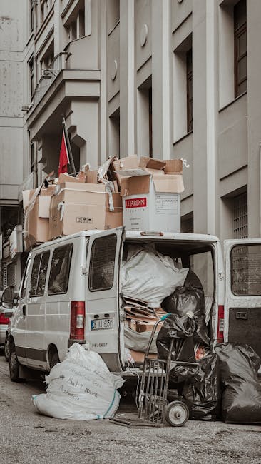 A white van with its rear doors open is parked on a narrow urban street in front of a multi-storey building with a beige facade featuring multiple windows and architectural details. The van is loaded with various packing materials, including cardboard boxes, plastic-wrapped furniture, and large bags of items, some of which are stacked on top of each other. Several cardboard boxes, some labeled with shipping or delivery tags, are positioned in and around the vehicle, indicating an active home relocation or moving process. A small hand trolley is also visible next to the van, loaded with additional black plastic bags. The scene suggests the final stages of packing or the transition of furniture and belongings during a house move, with equipment such as blankets or straps possibly used for securing items during transport. This setting reflects a professional removal service working in a densely built residential area, designed to facilitate efficient furniture transport through narrow access points, consistent with services offered by Man and Van Ilford for removals.