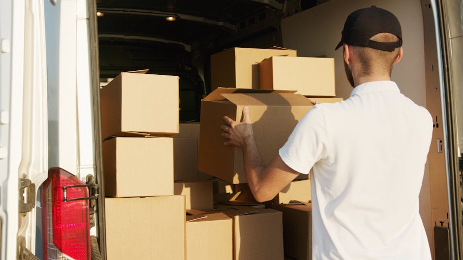 A man wearing a dark padded vest and blue long-sleeve shirt is standing outside near a white van, holding three cardboard boxes stacked together. The boxes are sealed with packing tape and have labels on them, indicating they are packed for home relocation or moving purposes. Inside the van, a person is visible, crouching near a stack of similar boxes and packing materials, suggesting they are involved in packing and loading processes. The scene takes place on a paved area adjacent to a modern building with large glass doors and metal framing, with natural daylight illuminating the scene. The image captures the loading process typical of furniture transport and packing during residential moves, reflecting the logistical steps undertaken by removals services such as Man and Van Ilford, especially when navigating narrow access points for flats or apartments.