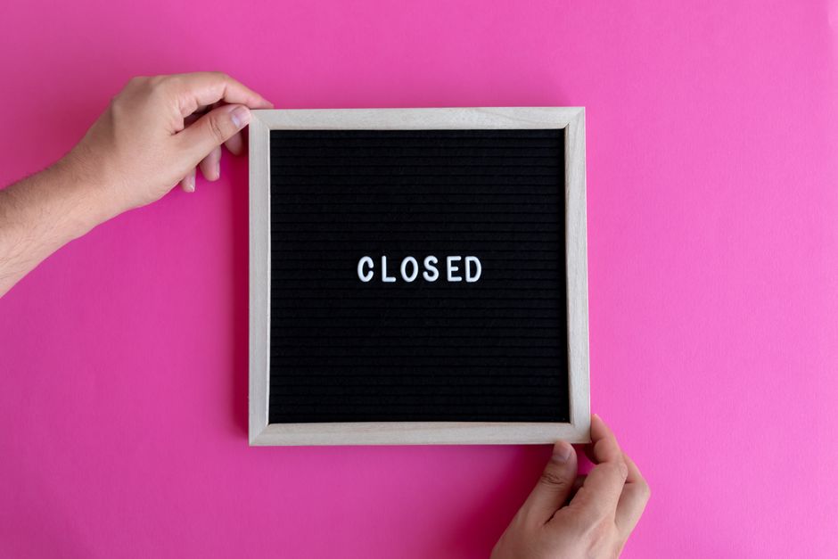 A person holds a small square black letterboard with a light wooden frame, displaying the word 'CLOSED' in white uppercase letters, against a bright pink background. One hand grips the top left corner