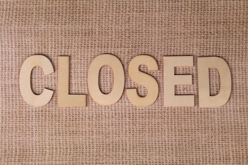 A person holds a small square black letterboard with a light wooden frame, displaying the word 'CLOSED' in white uppercase letters, against a bright pink background. One hand grips the top left corner