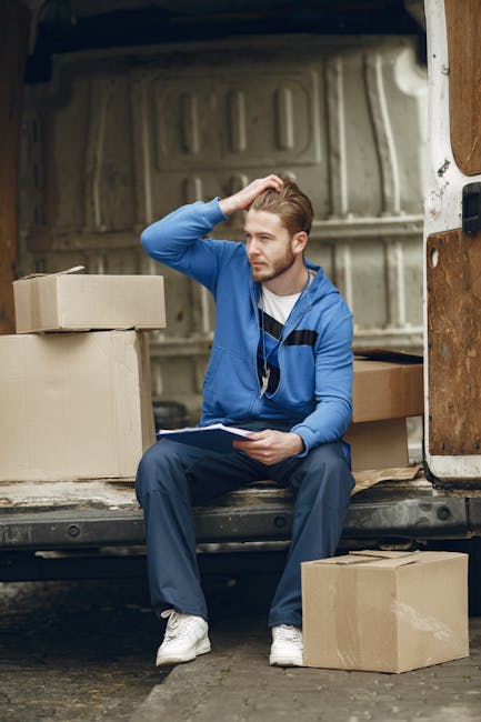 A young man with light brown hair, wearing a blue hoodie and navy trousers, is sitting on the edge of an open moving van's loading area, surrounded by sealed cardboard boxes of various sizes, some stacked and others placed on the ground beside him. He is holding a clipboard and appears to be reviewing inventory or planning the next step in a home relocation process. The interior of the van features wooden panels and a metal frame, with additional packing materials such as plastic wrap and blankets visible in the background. The scene is set outdoors on an overcast day, with natural light illuminating the workspace, and the overall environment suggests an organized loading or unloading operation, typical of furniture transport and packing during a house move managed by [COMPANY_NAME].
