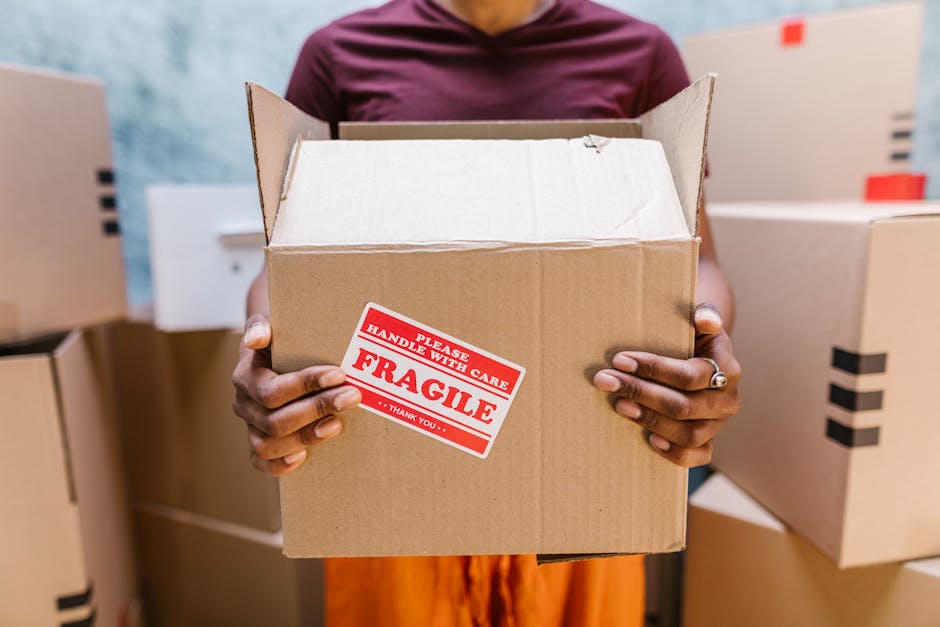 A person holding an open cardboard box with a red and white 'Fragile' sticker that reads 'Please Handle With Care' in a storage or packing facility, surrounded by several other closed boxes of various sizes. The individual’s hands, with a ring visible on one finger, grip the box firmly while standing in front of shelves stacked with additional boxes. The background includes soft lighting and a slightly blurred environment indicative of a professional moving or packing setting, with some boxes positioned on the floor and on shelving units. This scene captures the careful handling involved in home relocation and furniture transport, emphasizing proper packaging practices to ensure safe moving and packing processes, as often managed by services such as Man and Van Ilford.