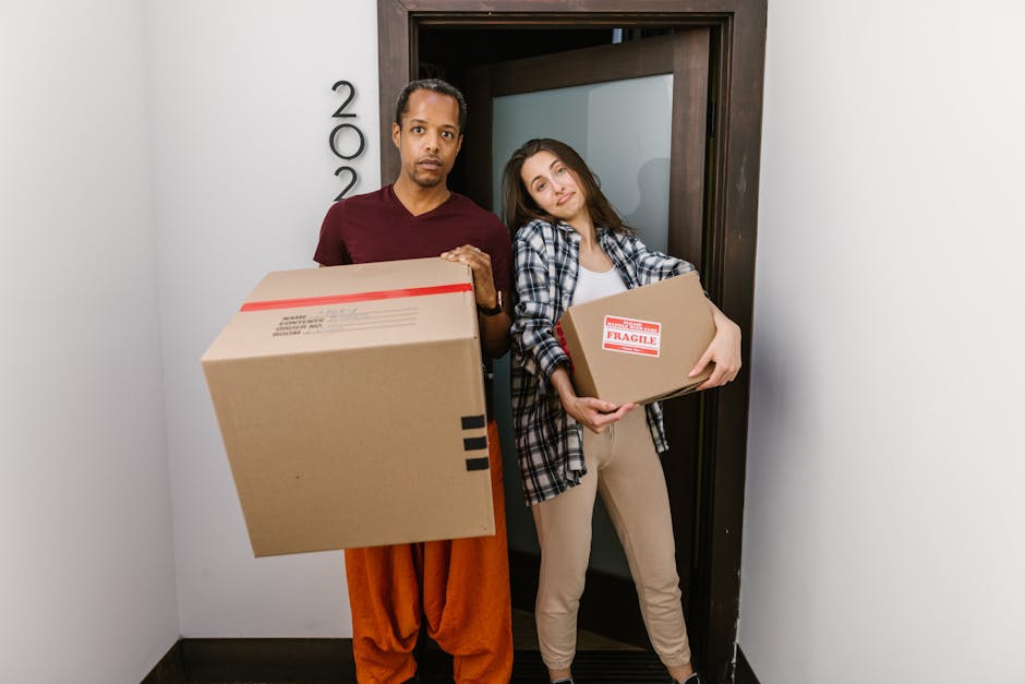 A man and a woman stand inside a doorway of a property, each holding cardboard boxes used for packing during home relocation. The man, on the left, is wearing a maroon t-shirt and orange pants, with a serious expression, holding a large box with red and black packing labels. The woman, on the right, is dressed in a plaid shirt over a white top and beige trousers, leaning slightly with a smile, holding a smaller box labeled 'Fragile.' Behind them, the door frame is dark wood, and the wall on the left displays the house number '200.' The background seen through the open doorway shows a frosted glass panel, indicating interior space readiness for moving or packing activities. The scene captures the typical process of furniture transport and packing for a house removal, with visible packing materials, boxes, and equipment suggesting the ongoing loading or unpacking process, which aligns with logistical aspects of professional removals services like those offered by Man and Van Ilford.
